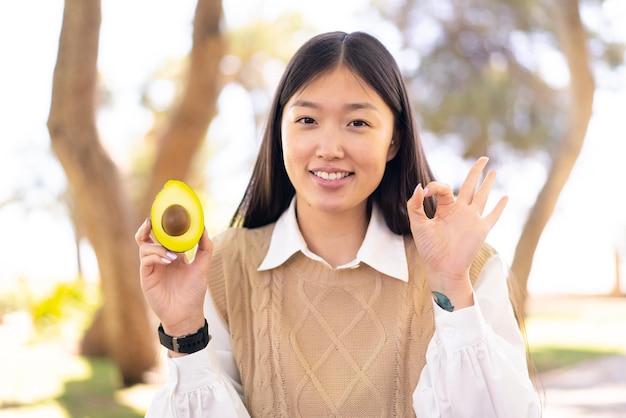 Linda mulher chinesa segurando um abacate ao ar livre mostrando sinal de ok com os dedos