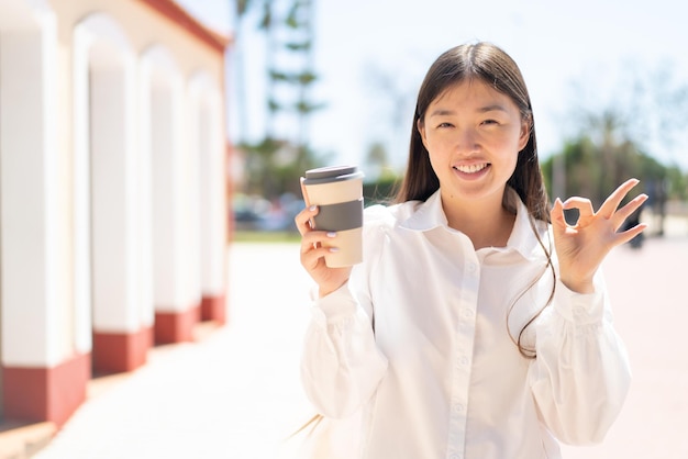 Linda mulher chinesa segurando um café para viagem ao ar livre mostrando sinal de ok com os dedos