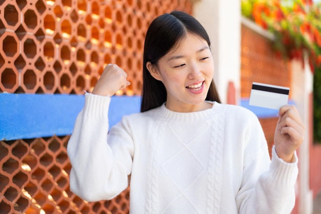 Linda mulher chinesa segurando um cartão de crédito ao ar livre comemorando uma vitória