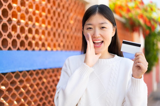 Linda mulher chinesa segurando um cartão de crédito ao ar livre gritando com a boca aberta