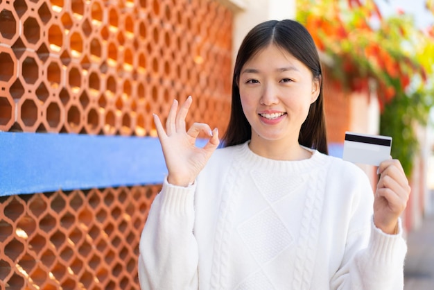 Linda mulher chinesa segurando um cartão de crédito ao ar livre mostrando sinal de ok com os dedos