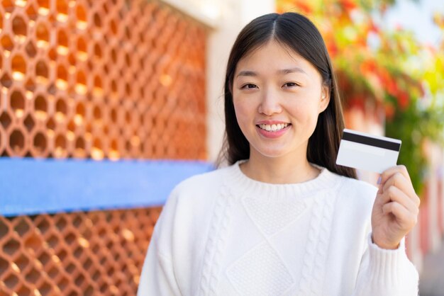 Linda mulher chinesa segurando um cartão de crédito ao ar livre sorrindo muito