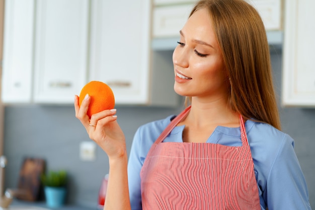 Linda mulher com avental vermelho segurando laranjas maduras enquanto está na cozinha