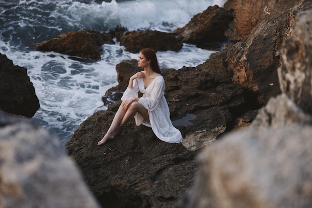 Linda mulher com cabelo molhado em vestido branco senta-se em pedras de ondas inalteradas