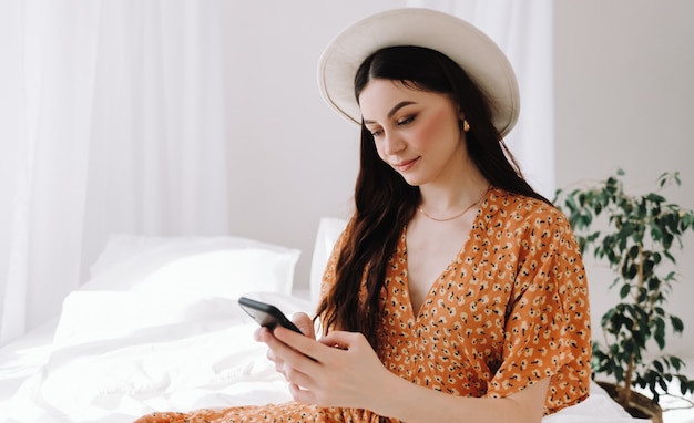 Linda mulher com chapéu branco sentada na cama e usando o smartphone em uma manhã ensolarada