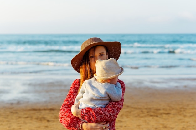 Linda mulher com chapéu e vestido vermelho segurando o filho na praia no pôr do sol de primavera na Espanha