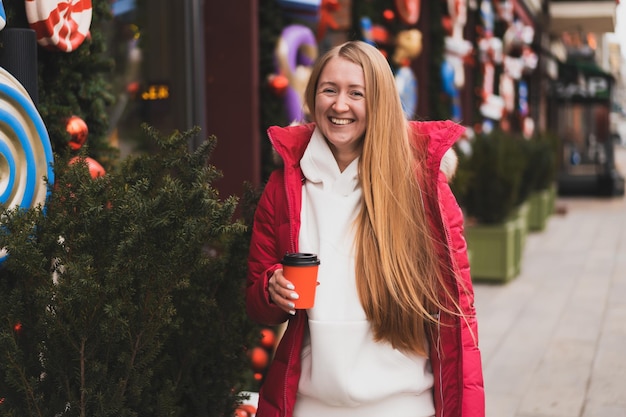 Linda mulher com roupas de inverno caminha pela rua decorada com decoração de ano novo e bebe café quente
