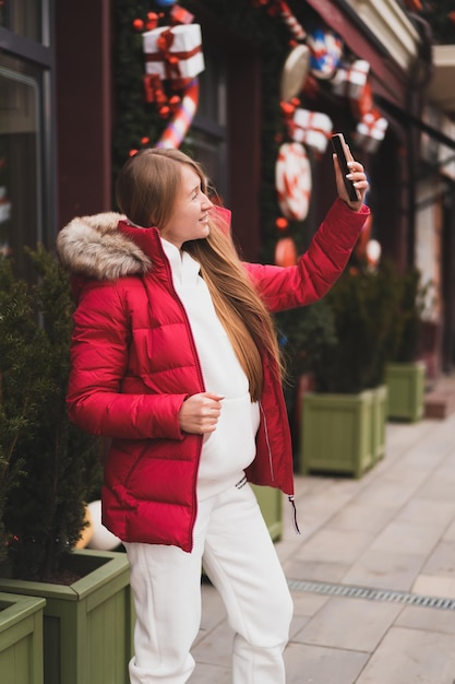 Linda mulher com roupas de inverno caminha pela rua decorada com decoração de ano novo e tira selfie no telefone