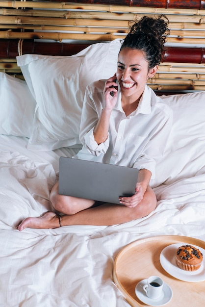 Foto linda mulher com telefone e laptop na cama