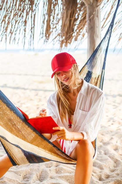 Linda mulher de boné vermelho escrevendo algo em seu planejador vermelho sentado em uma rede na praia Planejamento de vida ou trabalho em ambiente confortável