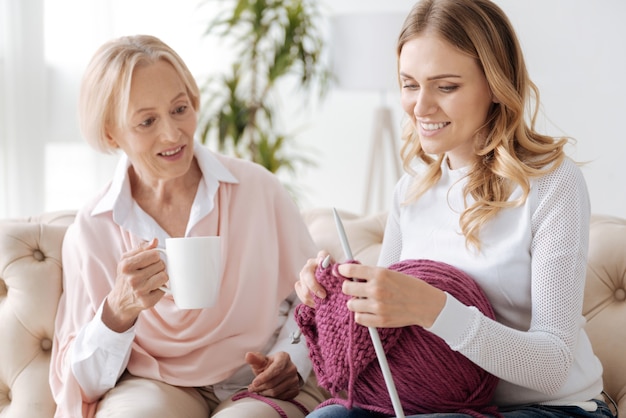 Linda mulher de cabelos dourados tricotando um lenço com fios roxos e segurando uma grande bola de fios no colo, enquanto sua mãe elegante segurando uma caneca de chá e olhando para seu tricô