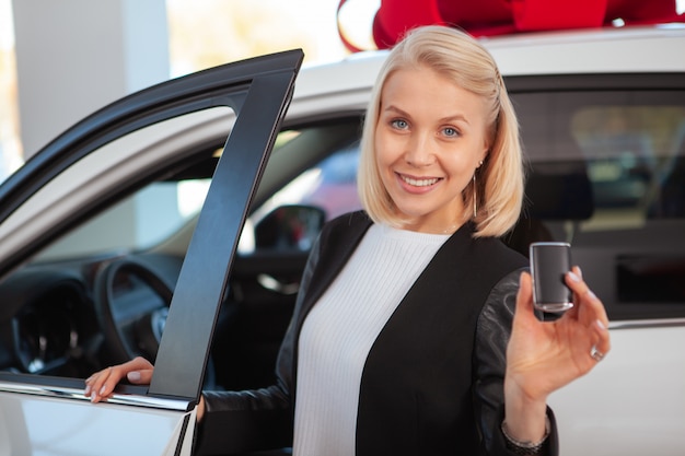 Linda mulher de cabelos loiro sorrindo, segurando a chave do carro na concessionária