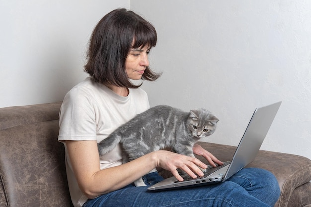 Linda mulher de meia idade com um gato fofo trabalha em casa em um laptop Mulher de meia idade trabalha remotamente Conceito de local de trabalho em casa com animais de estimação Animais de estimação fazem parte da família