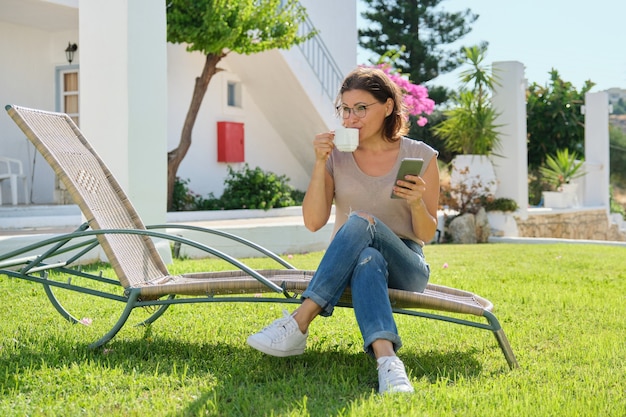 Linda mulher de meia idade descansando lendo, bebendo café conversando. mulher sentada na espreguiçadeira do jardim no gramado perto da casa. estilo de vida, lazer, tecnologia, beleza de pessoas maduras