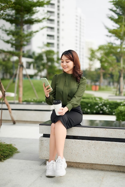 Linda mulher de negócios com roupa verde casual sentada no parque, segurando café e falando ao telefone