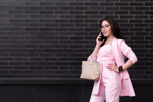 Linda mulher de negócios de óculos em um terno rosa com uma bolsa fica no contexto de uma parede de tijolos pretos falando em um telefone celular