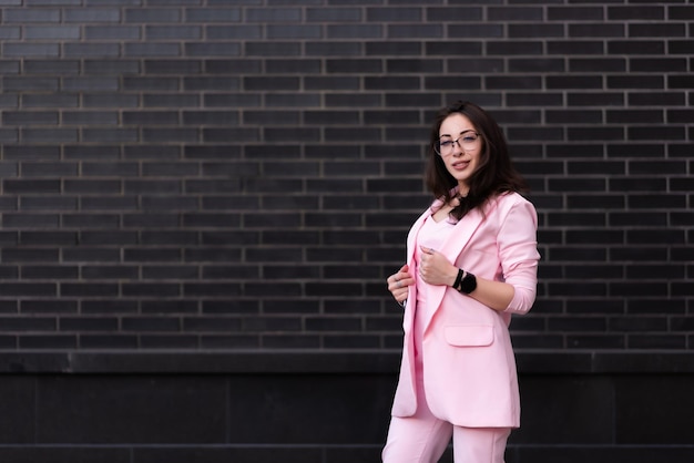 Linda mulher de negócios de óculos em um terno rosa fica no contexto de uma parede de tijolos pretos Posando e olhando para a câmera