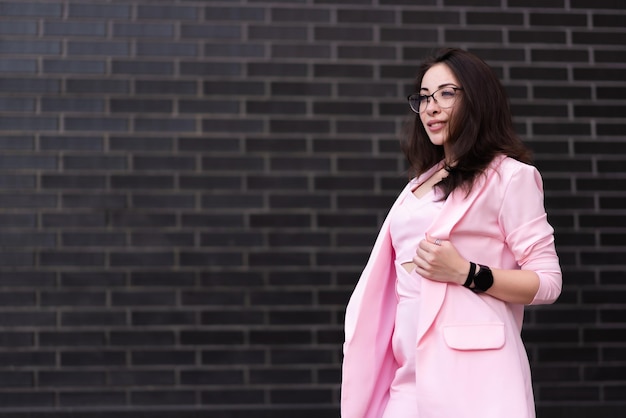Linda mulher de negócios de óculos em um terno rosa fica no contexto de uma parede de tijolos pretos Posando e olhando para longe