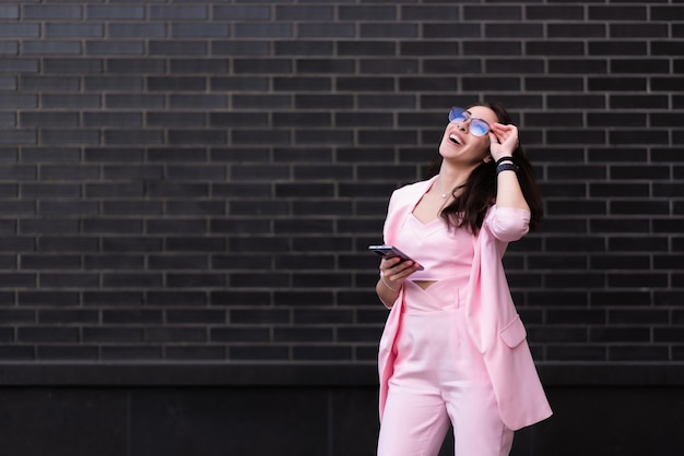 Linda mulher de negócios de óculos em um terno rosa fica no contexto de uma parede de tijolos pretos ri depois de ler uma mensagem em um telefone celular