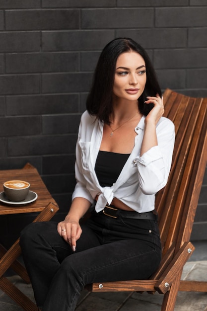 Linda mulher de negócios elegante com camisa branca senta-se e descansa na cadeira vintage de madeira e bebe café ao ar livre perto de uma parede de tijolo preto