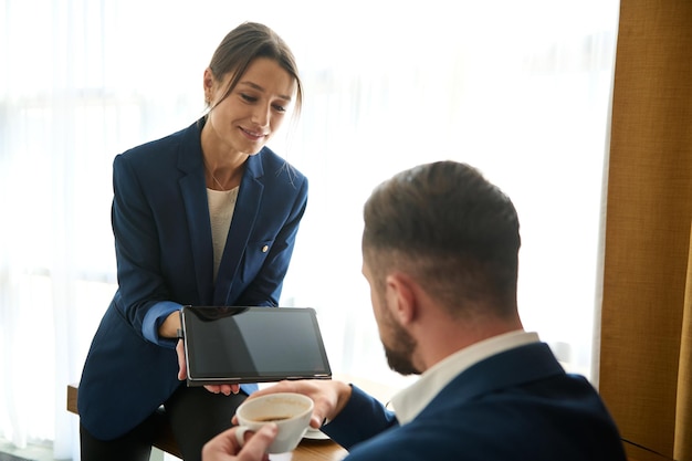 Foto linda mulher de negócios europeia de meia idade em um terno de negócio segurando um tablet com tela em branco e mostrando para seu chefe, sentado na frente dela e bebendo café. cope espaço para propaganda