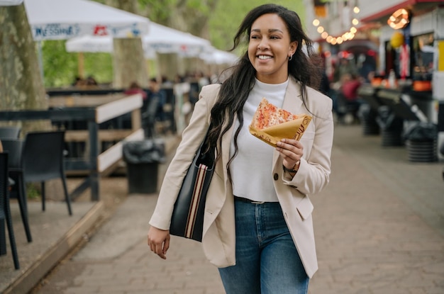 Linda mulher de raça mista sorridente segurando uma fatia de pizza olhando para longe, andando no mercado de rua