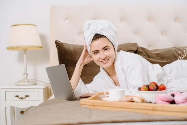 Linda mulher de roupão e toalha na cabeça usando um laptop na cama em casa