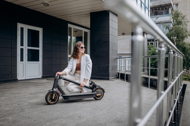 Linda mulher de terno branco dobrando sua eletro scooter depois de andar em pé no fundo do edifício moderno