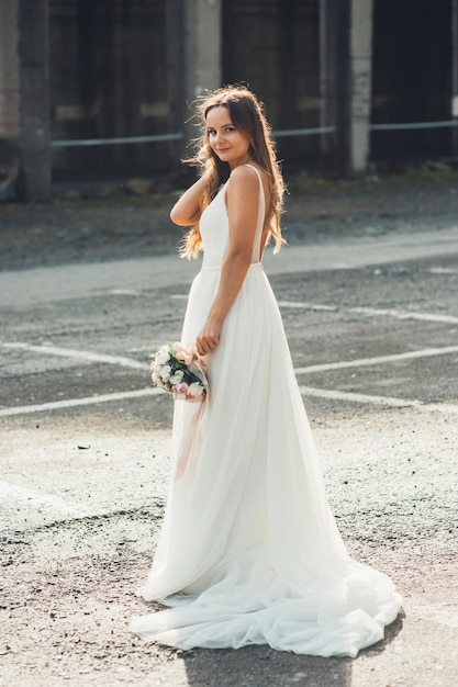 Linda mulher de vestido branco com buquê de flores posando ao ar livre na estrada e looki