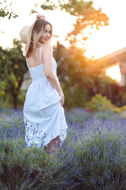 Linda mulher de vestido branco longo e chapéu fica em Lavender Meadow no conceito de verão Golden Hour