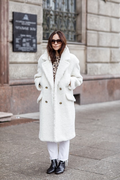 Linda mulher elegante andando na rua em um dia frio de inverno nevado. menina na moda vestindo blusa preta e calça e casaco de pele branca.