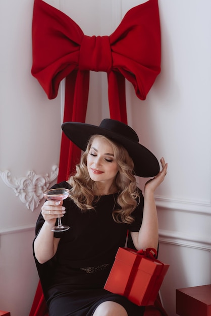 Linda mulher elegante em vestido festivo preto e chapéu com taça de champanhe e presentes de Ano Novo Foco seletivo suave