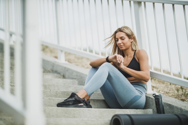 Linda mulher em forma está descansando antes de treinar duro nas escadas da ponte na cidade e olhando no smartwatch.