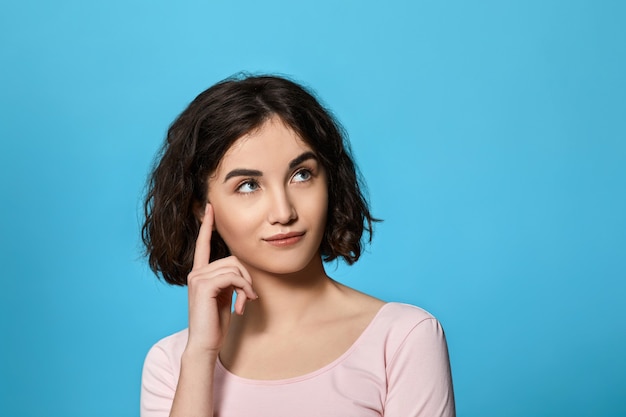 Linda mulher encaracolada morena pensando em algo sobre fundo azul. Emoções humanas, conceito de expressão facial.