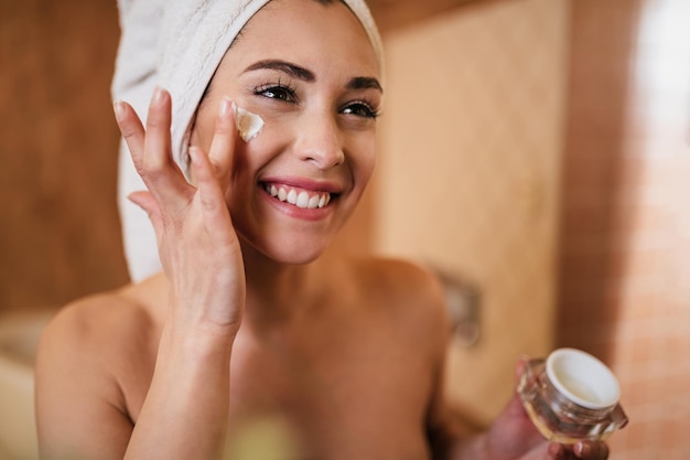 Linda mulher enrolada em toalha aplicando creme facial no banheiro