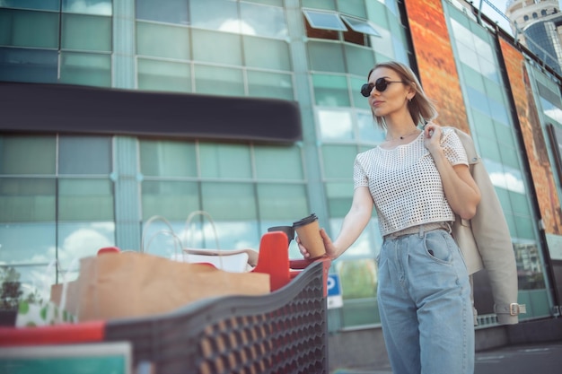 Linda mulher estilosa com carrinho de compras e compras bebe café em local urbano estilo de vida