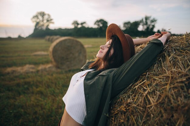 Linda mulher estilosa de chapéu relaxando no palheiro no campo noturno de verão Momento de tranquilidade