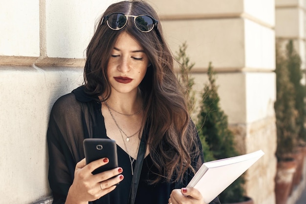 Linda mulher estilosa usando telefone e segurando revista na rua da cidade garota hipster feliz com roupa da moda mandando mensagens no smartphone na ensolarada cidade europeia