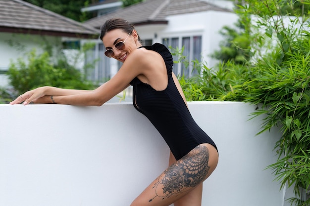 Linda mulher europeia em maiô preto posando do lado de fora da villa de férias forma perfeita bom corpo bronzeado usando óculos escuros marrons da moda