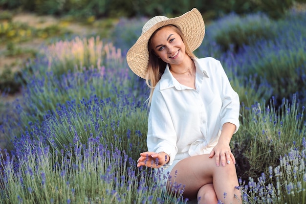 Linda mulher feliz curtindo a natureza em Lavender Meadow Jovem senta-se em camisa branca e chapéu