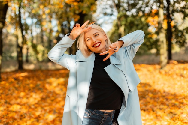 Linda mulher feliz e alegre com lábios vermelhos em uma camisola e um casaco azul da moda mostra o símbolo da paz e caminha em um parque dourado com folhagem de outono amarela. retrato feminino emocional engraçado