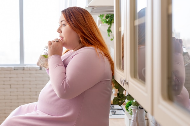 Linda mulher gordinha sentada na cozinha enquanto bebe um smoothie saudável