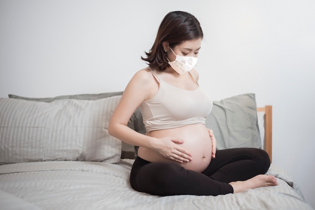 Linda mulher grávida asiática está usando máscara protetora em sua casa, conceito de proteção de Coronavírus