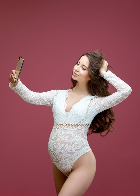 Linda mulher grávida com cabelos longos, vestida de lingerie de renda branca com um macacão fazendo uma selfie em um fundo rosa no estúdio.