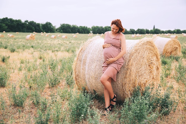 Linda mulher grávida de vestido na natureza Mulher segura as mãos na barriga no fundo do campo de trigo com palheiros no dia de verão Foto da expectativa de maternidade de gravidez Mãe esperando um bebê