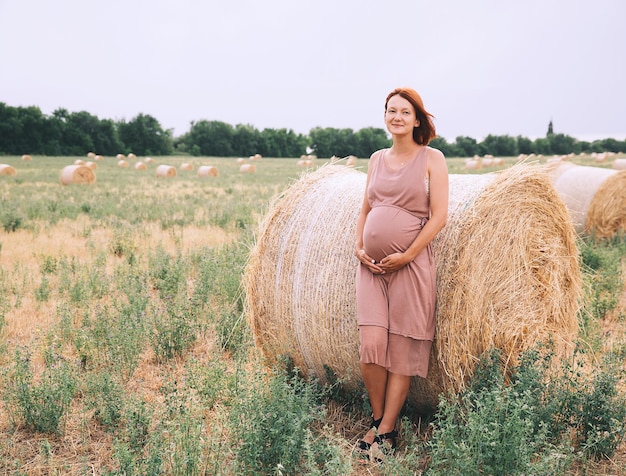 Linda mulher grávida de vestido na natureza Mulher segura as mãos na barriga no fundo do campo de trigo com palheiros no dia de verão Foto da expectativa de maternidade de gravidez Mãe esperando um bebê