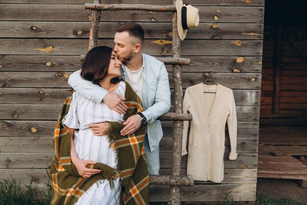 Linda mulher grávida e o marido posando perto de prédio de madeira. elegante casal feliz se amam. moda e estilo rústico.