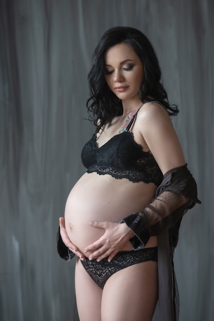 linda mulher grávida em cueca preta abraçando a barriga contra uma parede escura