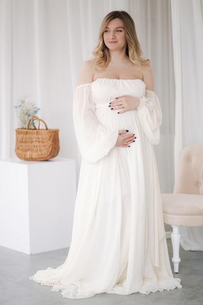 Linda mulher grávida em um elegante vestido branco posando para o fotógrafo  em um estúdio de fundo branco