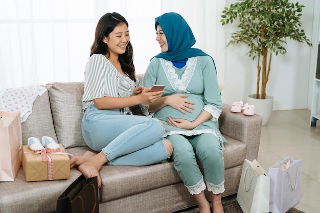 Linda mulher grávida muçulmana com lenço na cabeça e amigo usando telefone inteligente e sorrindo enquanto celebrava o chá de bebê. meninas sentadas no sofá com caixa de presente. duas senhoras compras online venda de verão
